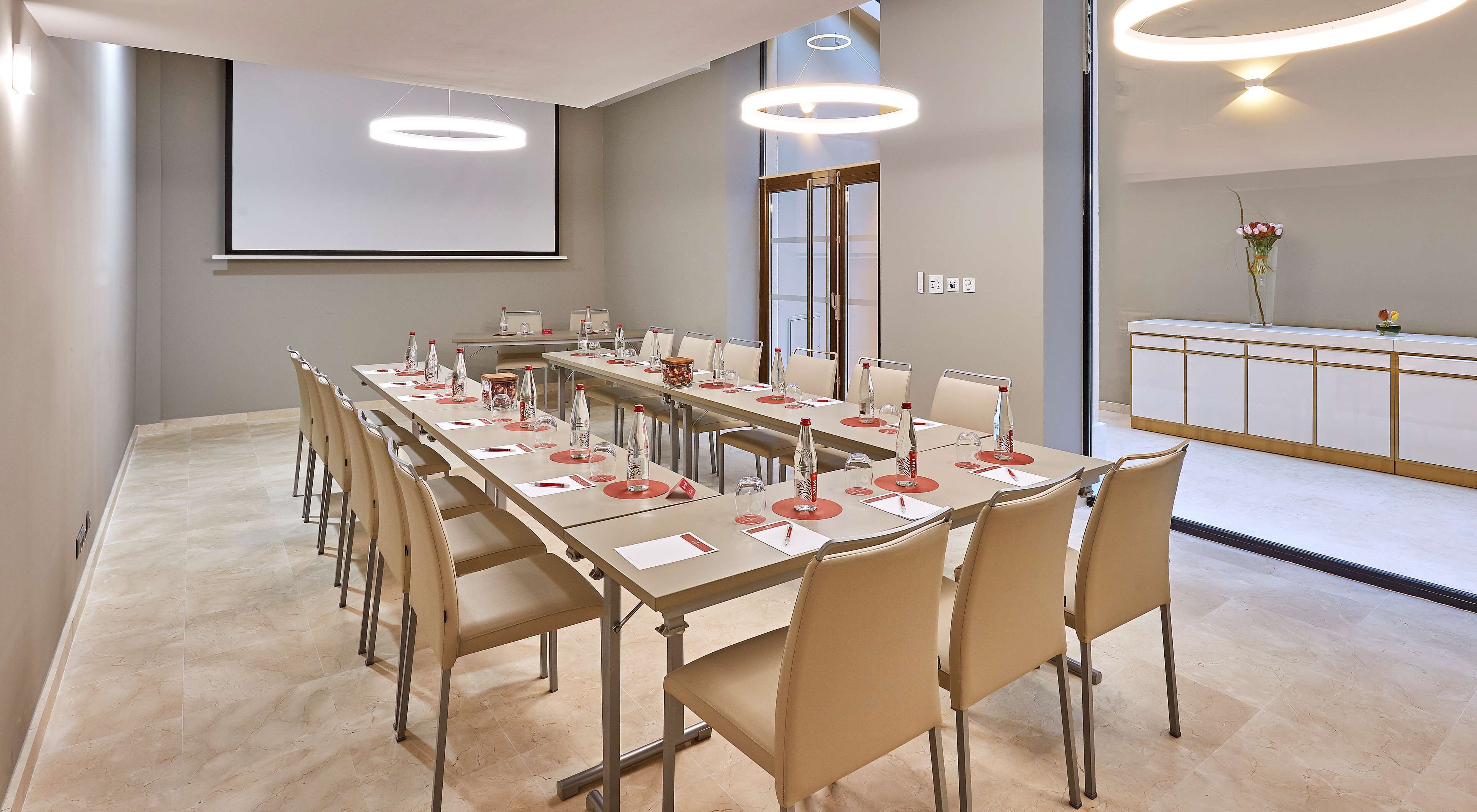 Un salón de conferencias con una larga mesa, sillas, una pantalla de proyector y una recepción con un jarrón de flores.