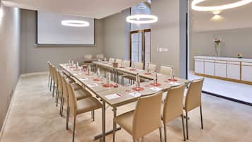 Un salón de conferencias con una larga mesa, sillas, una pantalla de proyector y una recepción con un jarrón de flores.