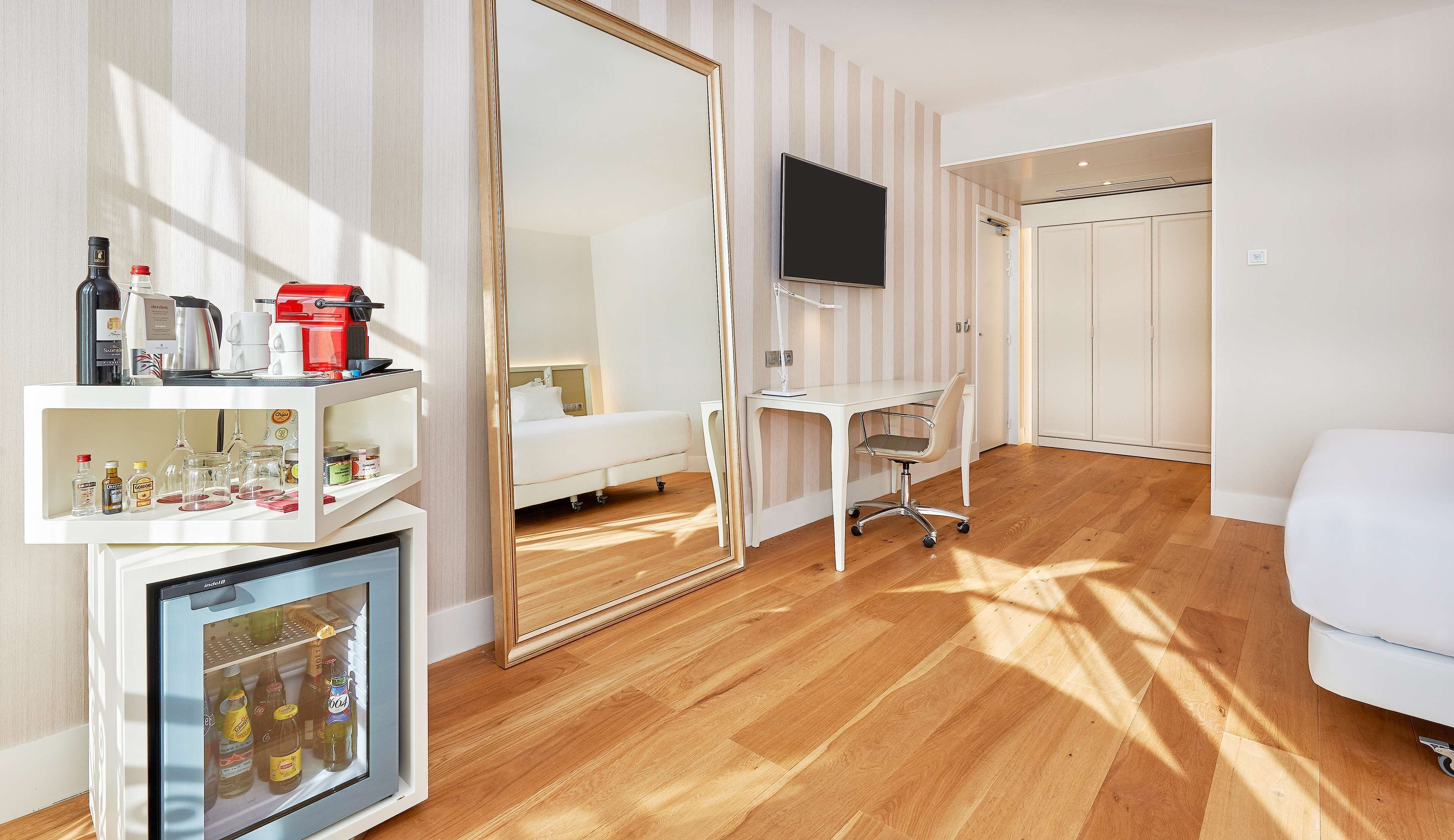 Habitación de hotel con piso de madera, una cama, un escritorio con silla, un televisor montado en la pared, un armario y un minibar con diversos artículos.