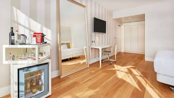 Habitación de hotel con piso de madera, una cama, un escritorio con silla, un televisor montado en la pared, un armario y un minibar con diversos artículos.