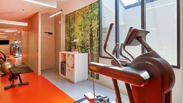 Un gimnasio moderno con equipamiento de ejercicio, una recepción y un amplio ventanal con vistas a árboles.
