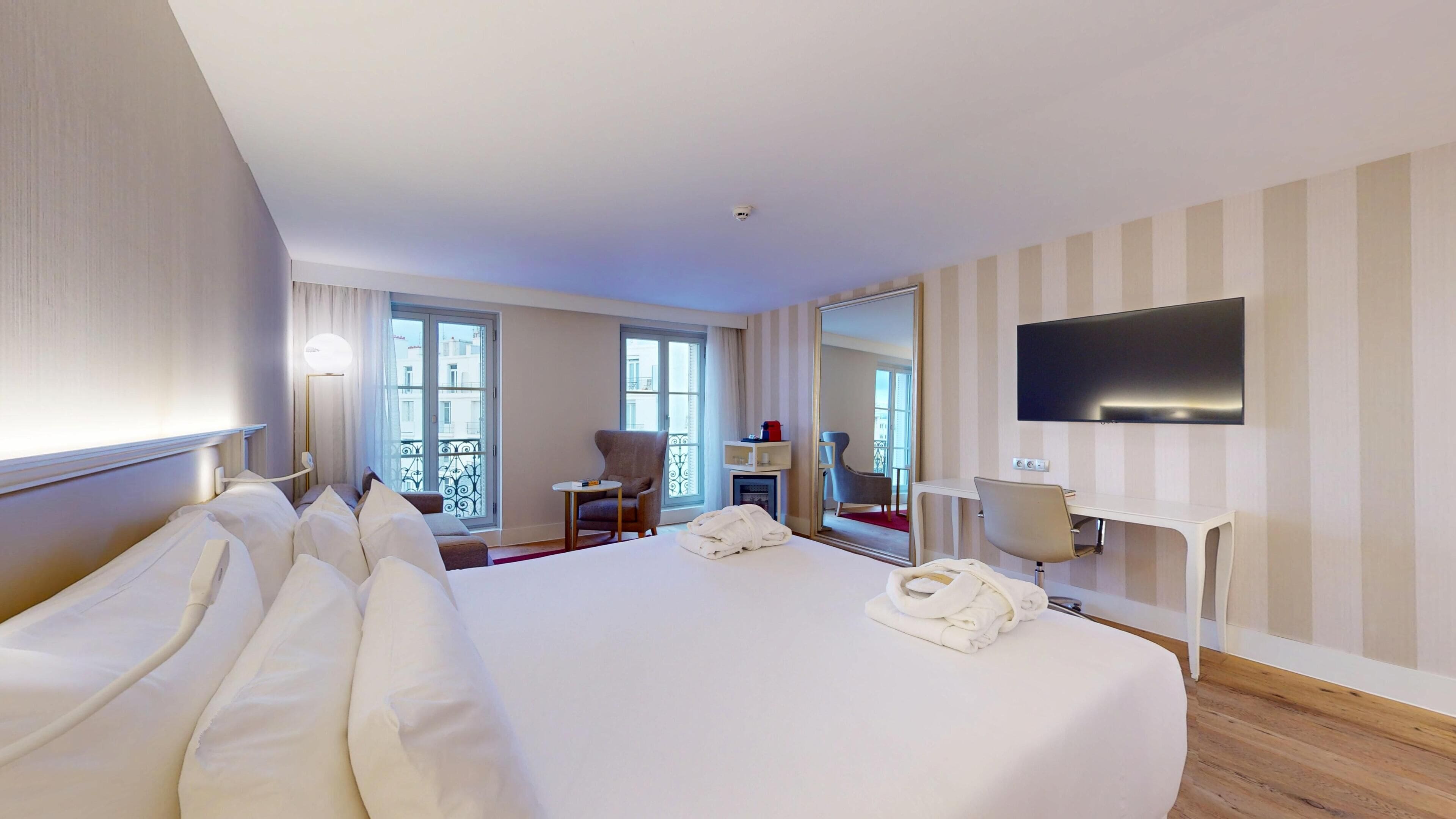 Habitación de hotel con una cama grande, un televisor montado en la pared, un escritorio con una silla y un balcón con vistas.