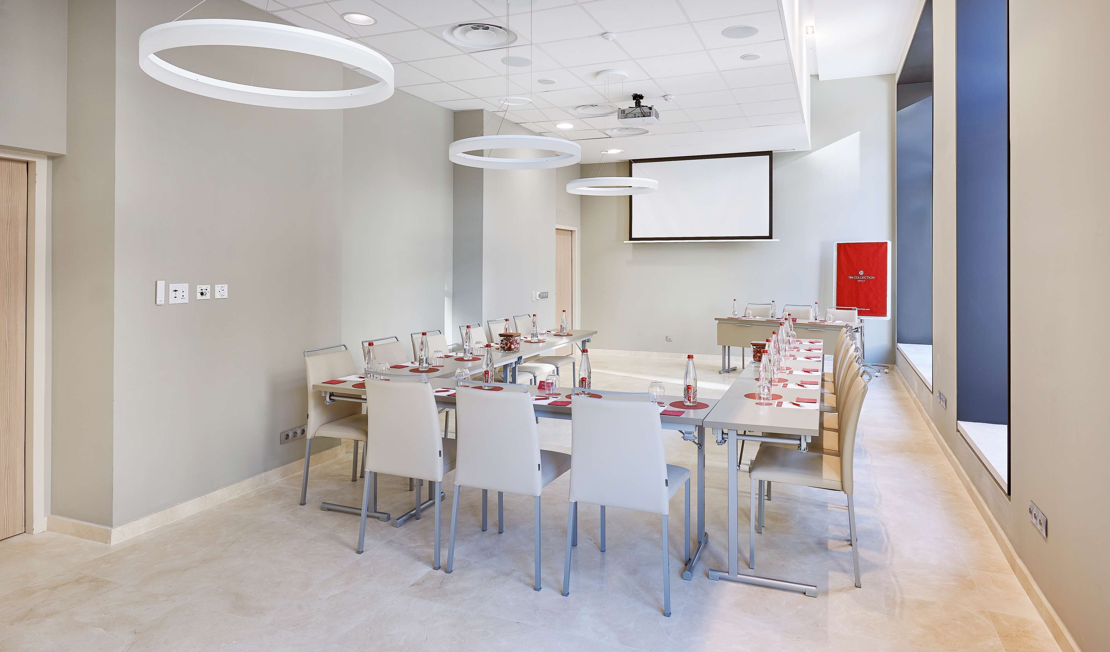Uma sala de conferência moderna com uma mesa comprida, cadeiras brancas e uma tela de projetor.