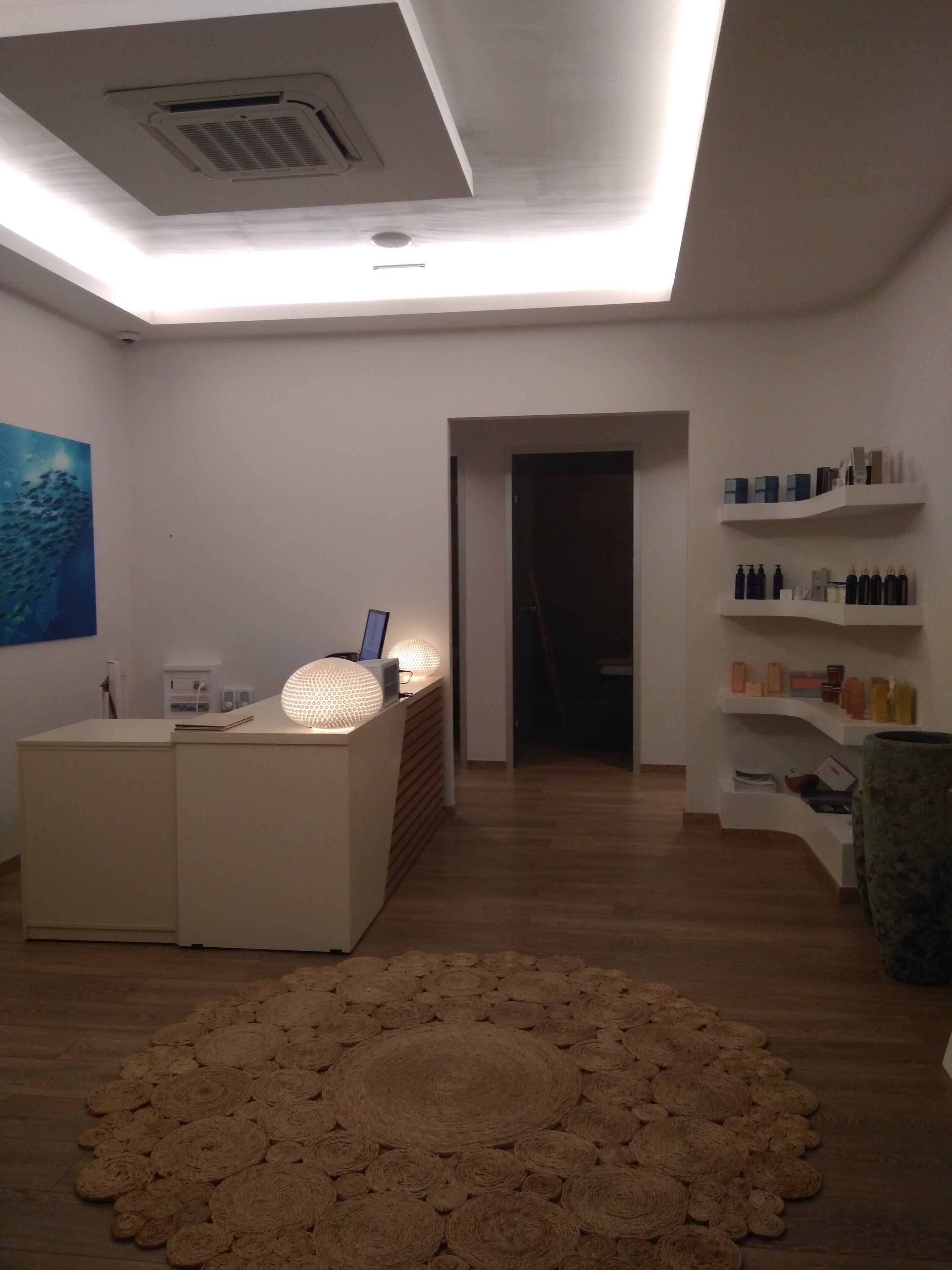 Un espace d’accueil moderne, doté d’un bureau, d’un ordinateur portable et d’un luminaire sphérique original. Des étagères contenant divers objets sont fixées au mur, et un grand tapis tissé recouvre le sol.