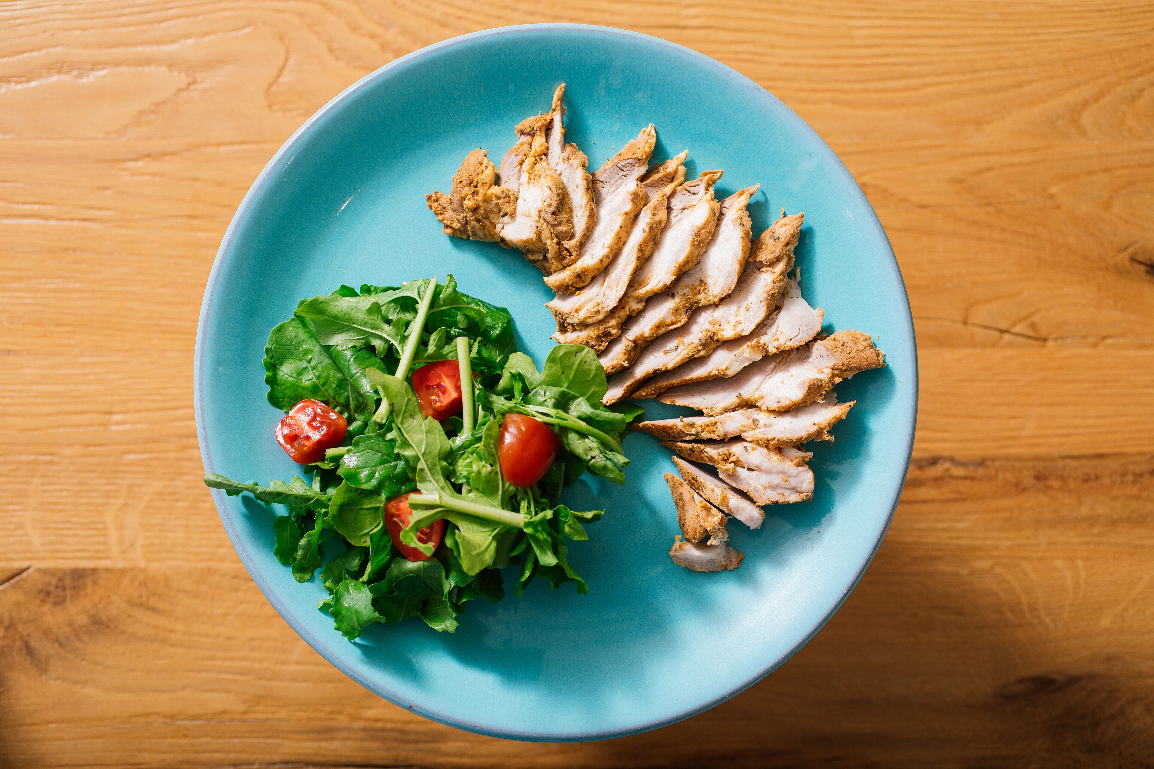 A blue plate with sliced chicken and a salad on a wooden table.