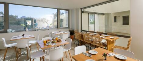 Un comedor moderno con ventanas amplias, sillas blancas, mesas de madera y vistas a la montaña.