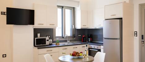 Une cuisine moderne avec des meubles blancs, un réfrigérateur en acier inoxydable, un téléviseur à écran plat fixé au-dessus du four à micro-ondes, et un coin repas avec deux chaises blanches et une table agrémentée d’un saladier de fruits et de verres à vin.