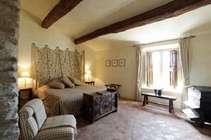 A cozy bedroom with a stone floor, a bed with a patterned bedspread, two bedside lamps, a bench, and a window with curtains. - La Verrière (Crestet)