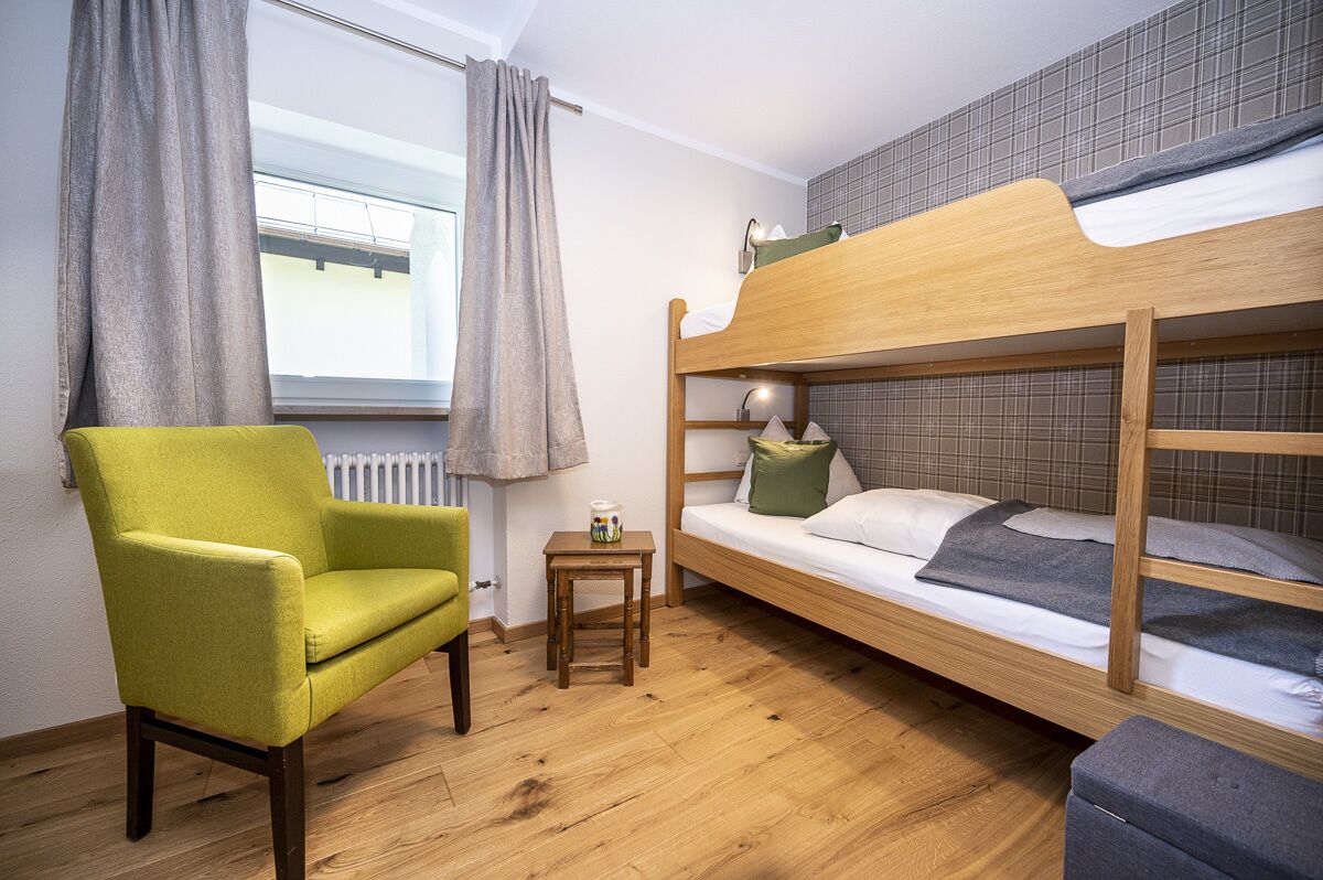 A room with a bunk bed, a green armchair, a small table, and a window with curtains.