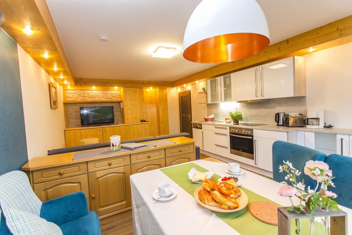 Eine moderne Küche mit Holzausstattung, einem Essbereich mit blauen Stühlen und einer Frühstücksecke mit Croissants und Obst.