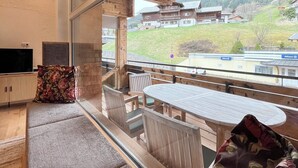Ein modernes Interieur mit Holzboden, einer Sitzecke mit Sofa und Sesseln, einem Esstisch und einem großen Fenster mit Blick auf ein Bergdorf.