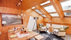 Ein Holzinnenraum mit einem für eine Mahlzeit gedeckten Esstisch, einer Couch und Bergblick durch ein Dachfenster.