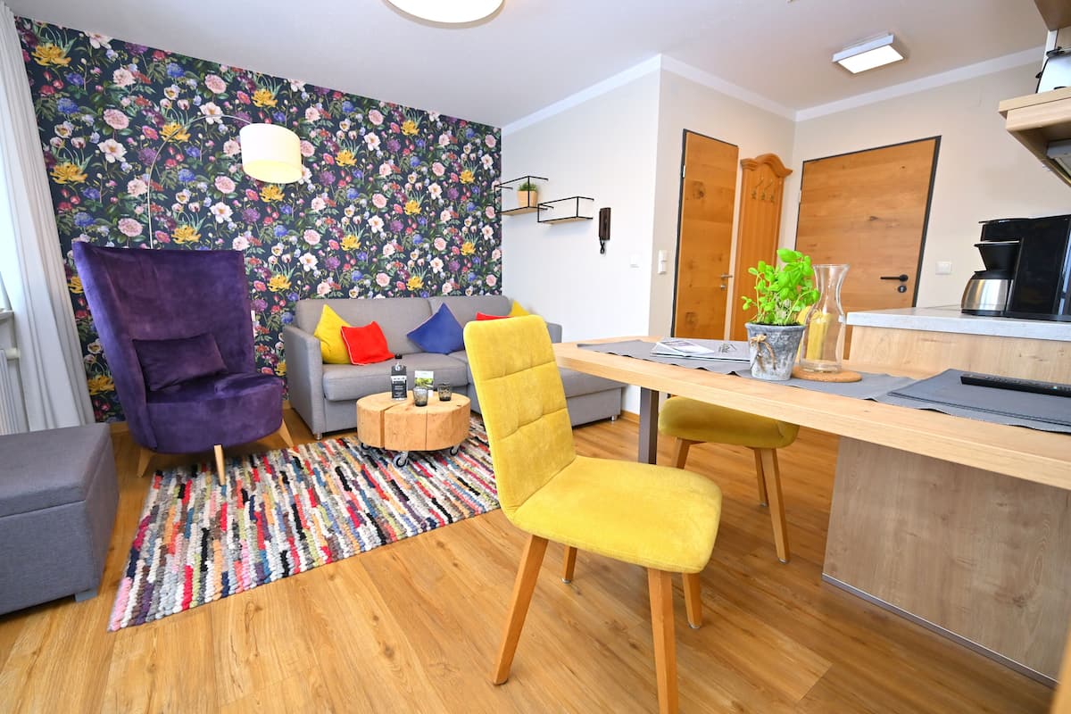 Ein modernes Wohnzimmer mit einer Couch, einem violetten Sessel und einem gelben Stuhl. Ein kleiner runder Tisch mit Pflanze und Lampe steht dort.