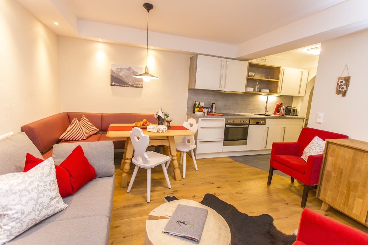 Ein modernes Wohnzimmer mit einer grauen Couch, roten und weißen Kissen, einem Holztisch und einem roten Sessel. Ein Essbereich mit Holztisch und weißen Stühlen grenzt an die Küche, die über weiße Schränke und einen Einbauofen verfügt.