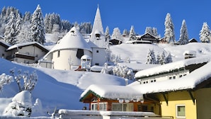 Ein verschneites Dorf mit traditioneller Architektur, darunter eine Kirche mit Spitzspitze und Häuser mit geneigten Dächern.
