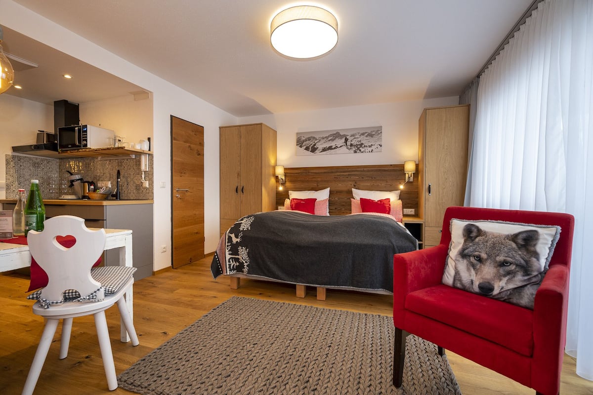 Ein modernes Schlafzimmer mit Bett, einem roten Sessel mit einem Wolfskissen, einem Holz-Kopfteil und einer Küchenzeile inklusive Mikrowelle und einer Flasche grüner Flüssigkeit.