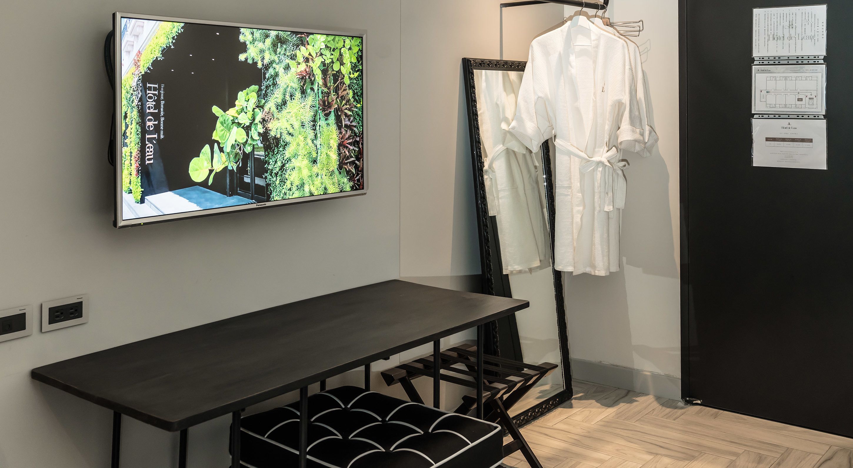 A hotel room with a large flat-screen TV mounted on the wall, a black metal rack with white robes, and a wooden table with black storage bins underneath.