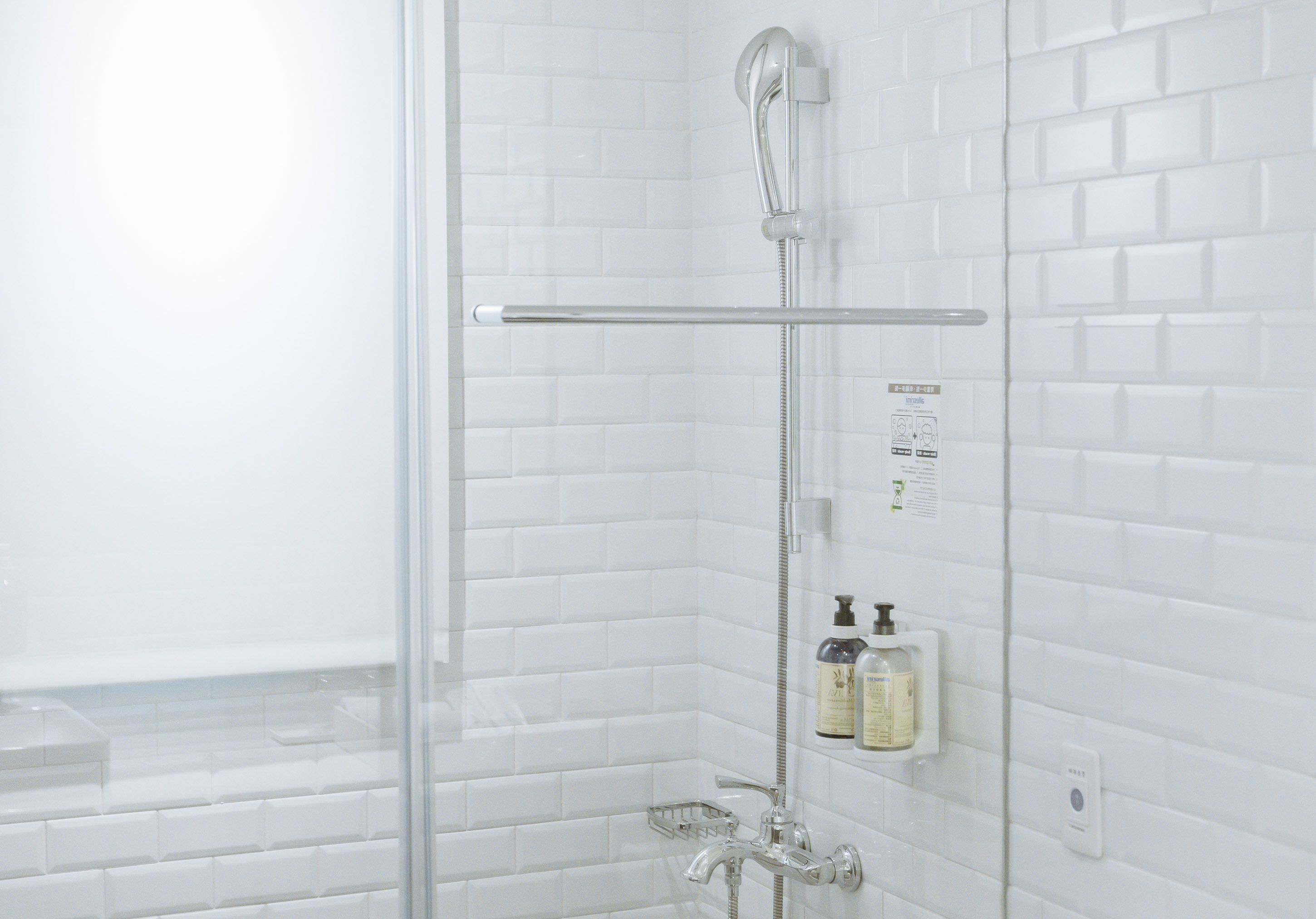 Un baño moderno con azulejos blancos tipo metro, mampara de vidrio, cabezal de ducha manual y dispensador de jabón en la pared.