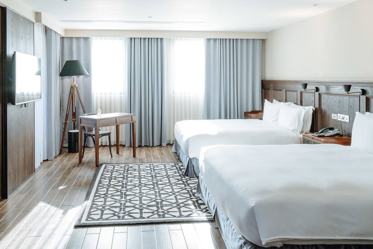Quarto de hotel com duas camas, uma escrivaninha, uma cadeira, um abajur e uma janela grande com cortinas.