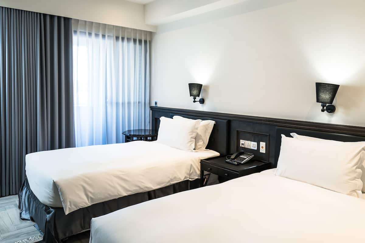 Quarto de hotel com duas camas, uma mesa de cabeceira com telefone e uma luminária na parede.