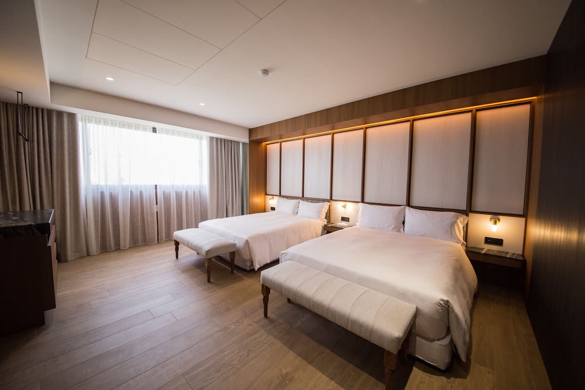 Quarto de hotel com duas camas, piso de madeira, uma janela grande com cortinas e luminárias de cabeceira.