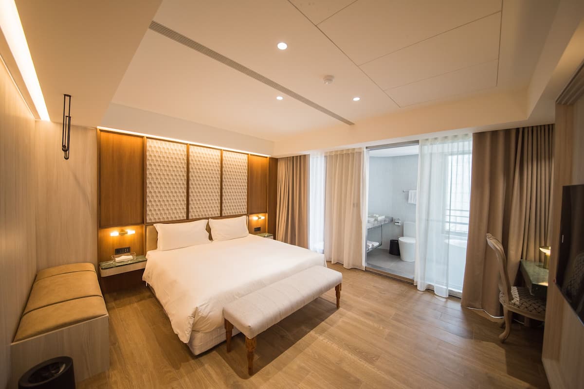 Quarto de hotel moderno com uma cama grande, mesas de cabeceira, um banco, uma TV e um banheiro visível por uma porta de vidro.