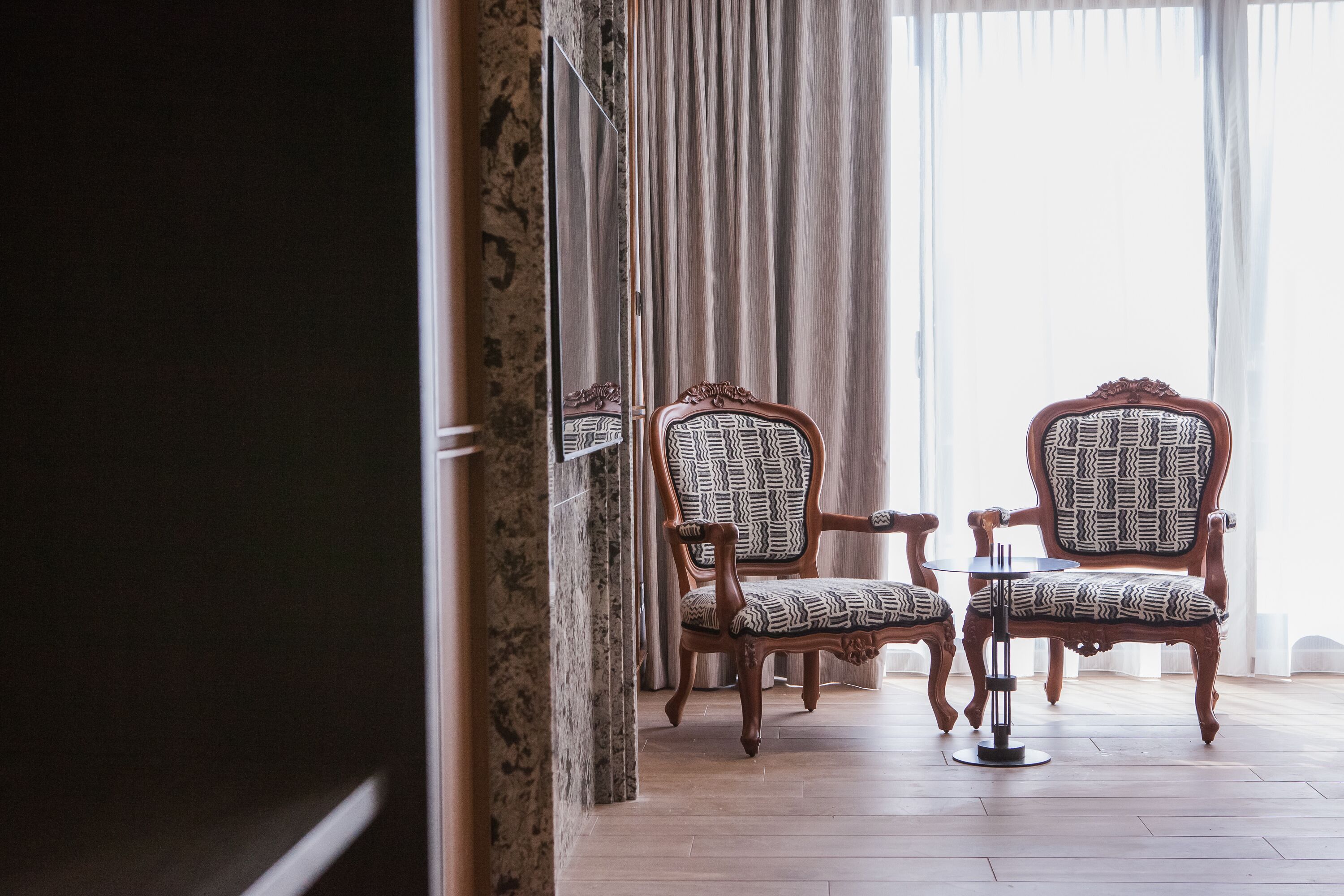 Una habitación con dos sillones estampados, una mesita redonda, una columna de mármol y un ventanal con cortinas.
