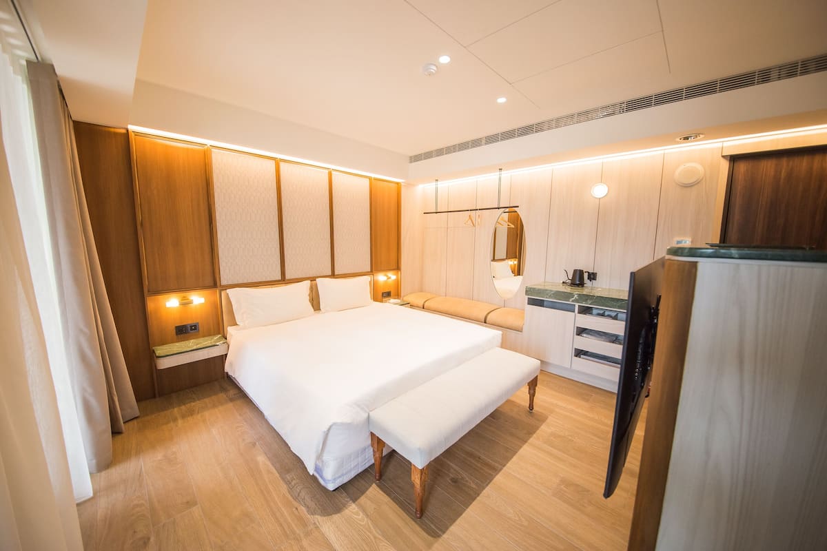 Quarto de hotel moderno com uma cama grande, mesas de cabeceira, um banco, um espelho e uma cozinha compacta.