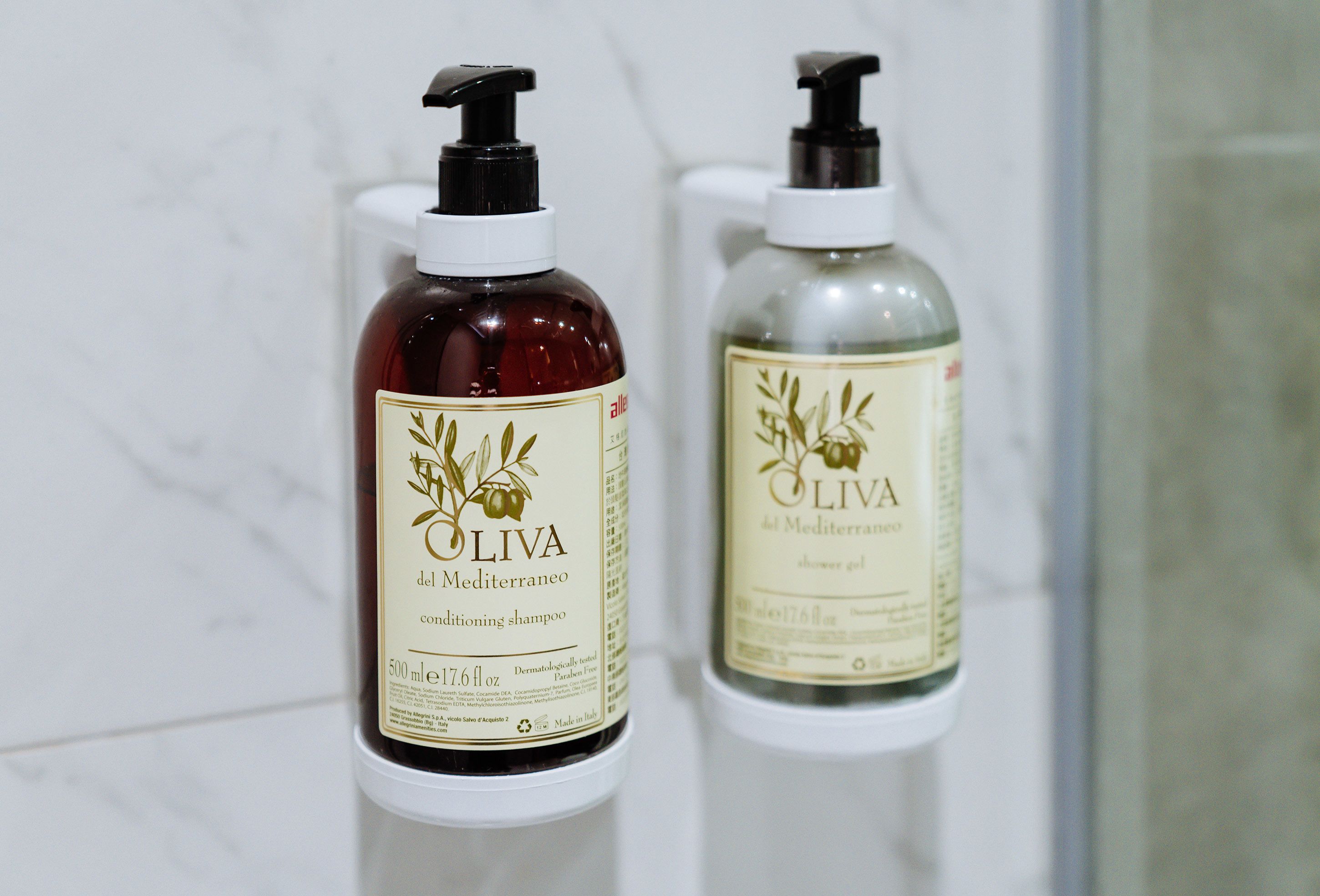 Dos botellas de champú y gel de ducha acondicionantes Oliva del Mediterraneo sobre un estante blanco.