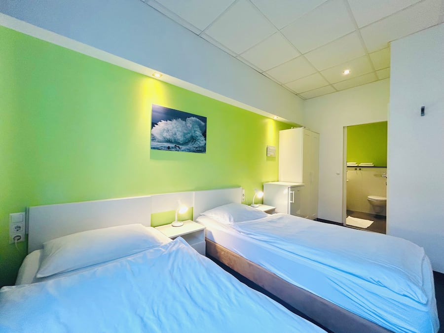 A hotel room with two beds, a green wall, a painting of a wave, and a bathroom visible through an open door.