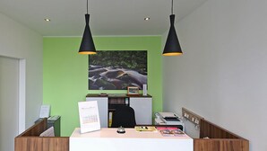 A reception desk with a wooden finish, two pendant lights, a framed picture of a river, and a small reception area with a desk, chair, and a printer. - Montagehotel Business & City (Linz)