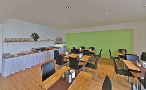 A dining area with multiple tables and chairs, a buffet setup, and a green accent wall. - Montagehotel Business & City (Linz)