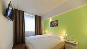 A double bed with white linens, a bedside table with a lamp, a TV mounted on the wall, and a window with curtains. - Montagehotel Business & City (Linz)