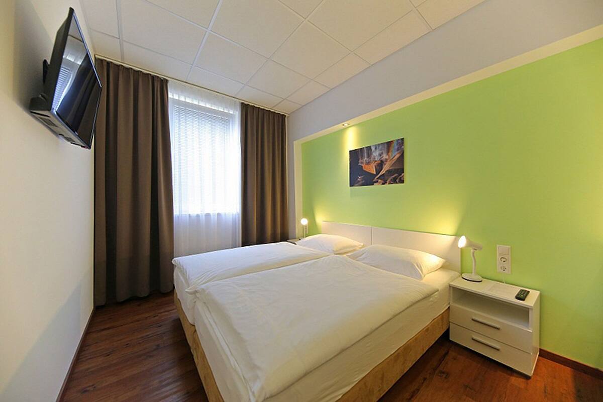Cama doble con sábanas blancas, mesita de noche con lámpara, televisor montado en la pared y ventana con cortinas.