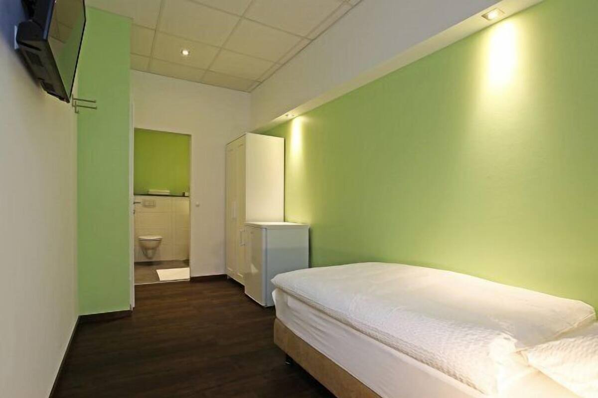 Un dormitorio con una cama individual, una mesita de noche pequeña y un espejo en la pared. Hay una puerta que da a un baño con inodoro y lavamanos.
