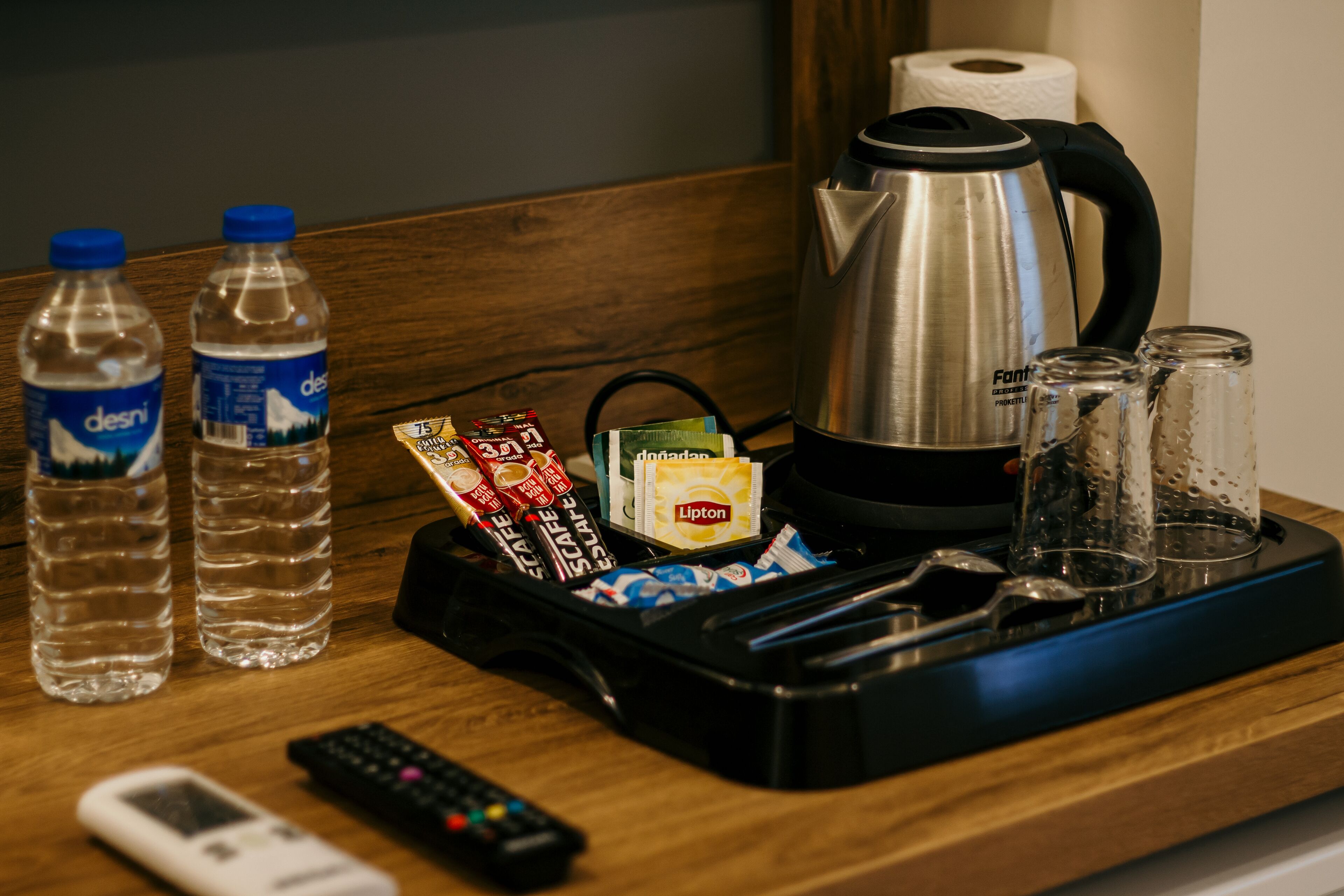 A tray with a kettle, water bottles, Lipton tea packets, and a remote control on a wooden surface.