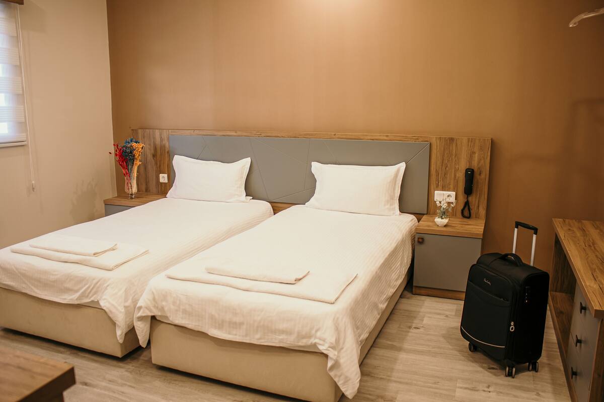 A hotel room with two beds, a wooden headboard, a nightstand with a phone, a vase with flowers, and a suitcase.