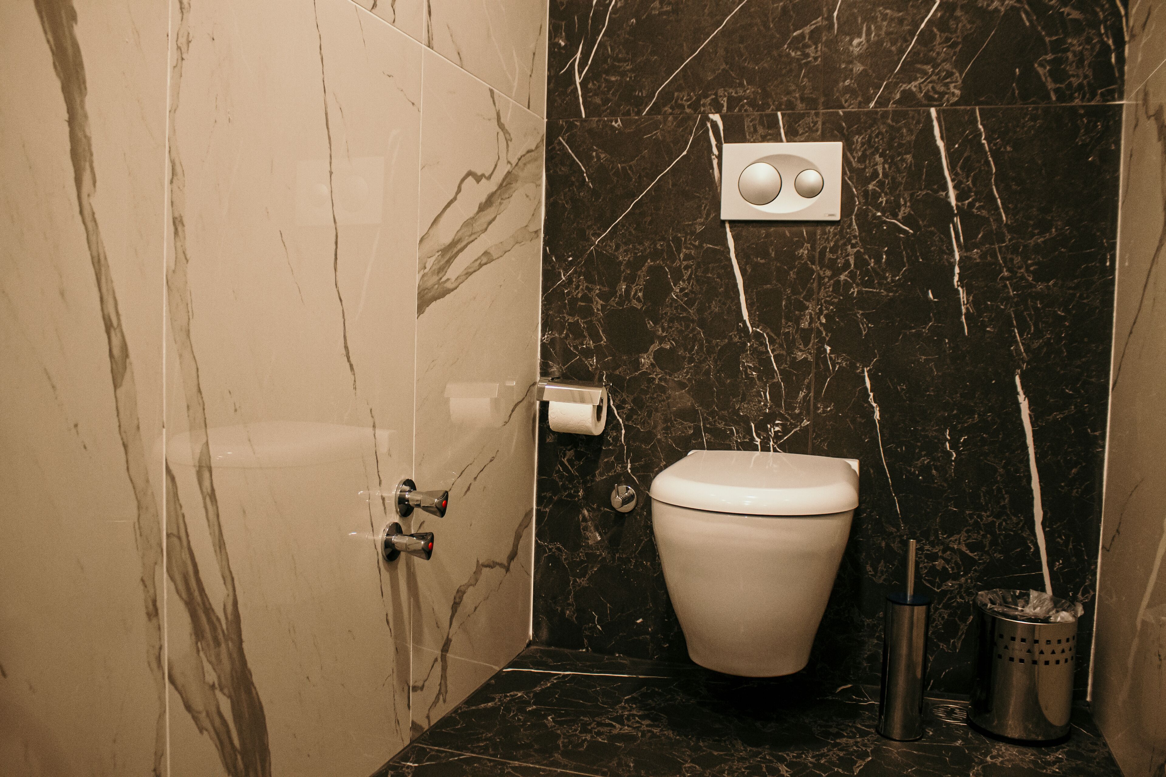 A bathroom with a toilet, marble tiles, a marble wall, a paper towel dispenser, and a trash can.