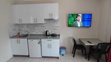 Una cocina compacta con armarios blancos, un refrigerador pequeño, una cafetera y un televisor en la pared.
