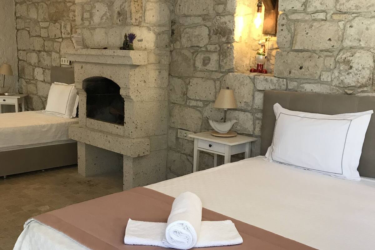 A room with a stone wall, a fireplace, a bed with white linens, and a nightstand with a lamp.