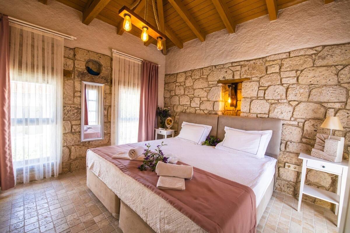 A bedroom with a stone wall, a bed with white bedding, a nightstand with a lamp, and a window with curtains.