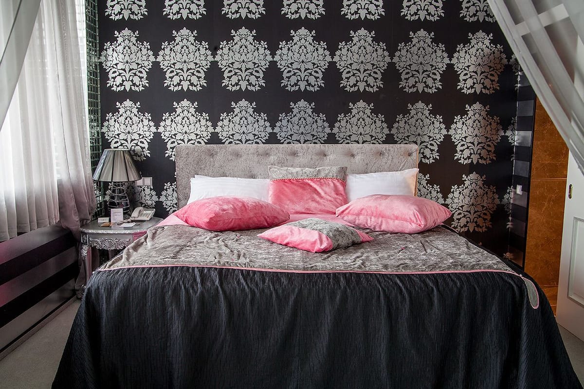 A bedroom with a tufted headboard, a bed with a dark grey and pink bedspread, and a patterned wallpaper.