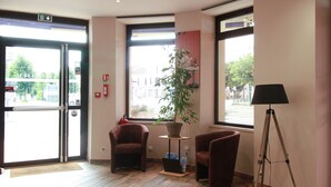 Un'hall d'albergo con pavimento in legno, due poltrone, una lampada da terra, una pianta in vaso e un'ampia finestra con vista sugli edifici esterni.