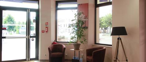 Un vestíbulo de hotel con piso de madera, dos sillones, una lámpara de pie, una planta en maceta y un amplio ventanal con vistas a edificios.