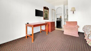 A hotel room with a wooden desk, a dresser, a pink armchair, a television, a mirror, and a lamp. - Capitol Inn And Suites (Montgomery)