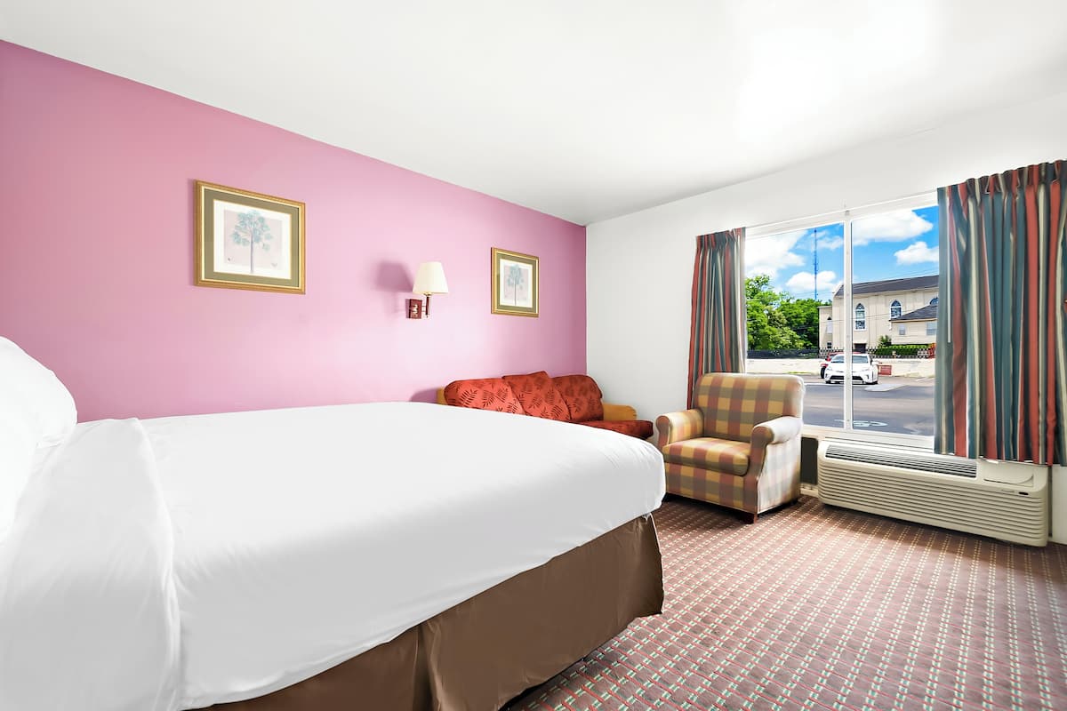 A hotel room with a large bed, a chair, a window with curtains, and two framed pictures on the wall.