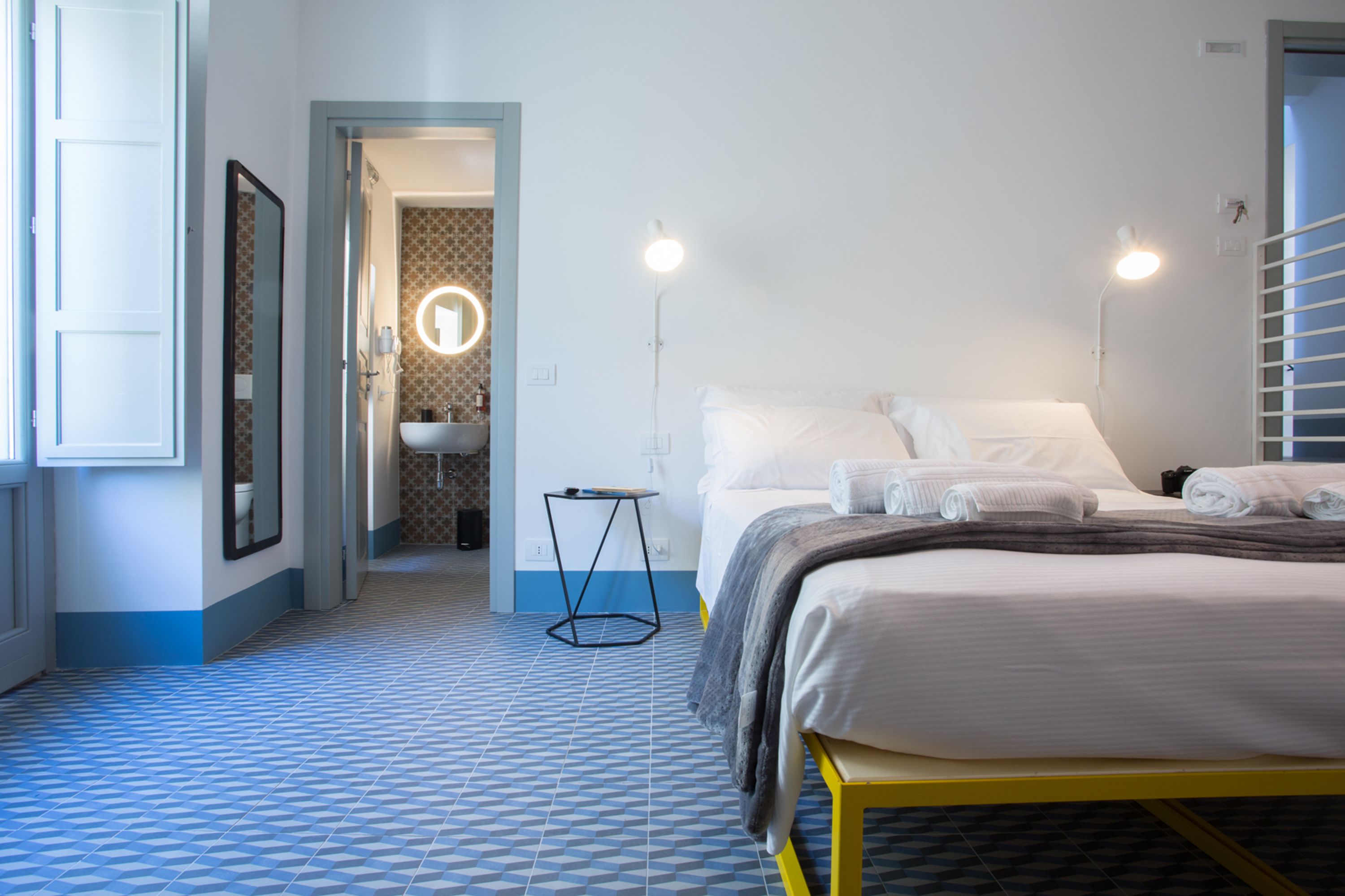Habitación de hotel con cama, lámparas de noche, una mesita y un baño visible a través de una puerta abierta.