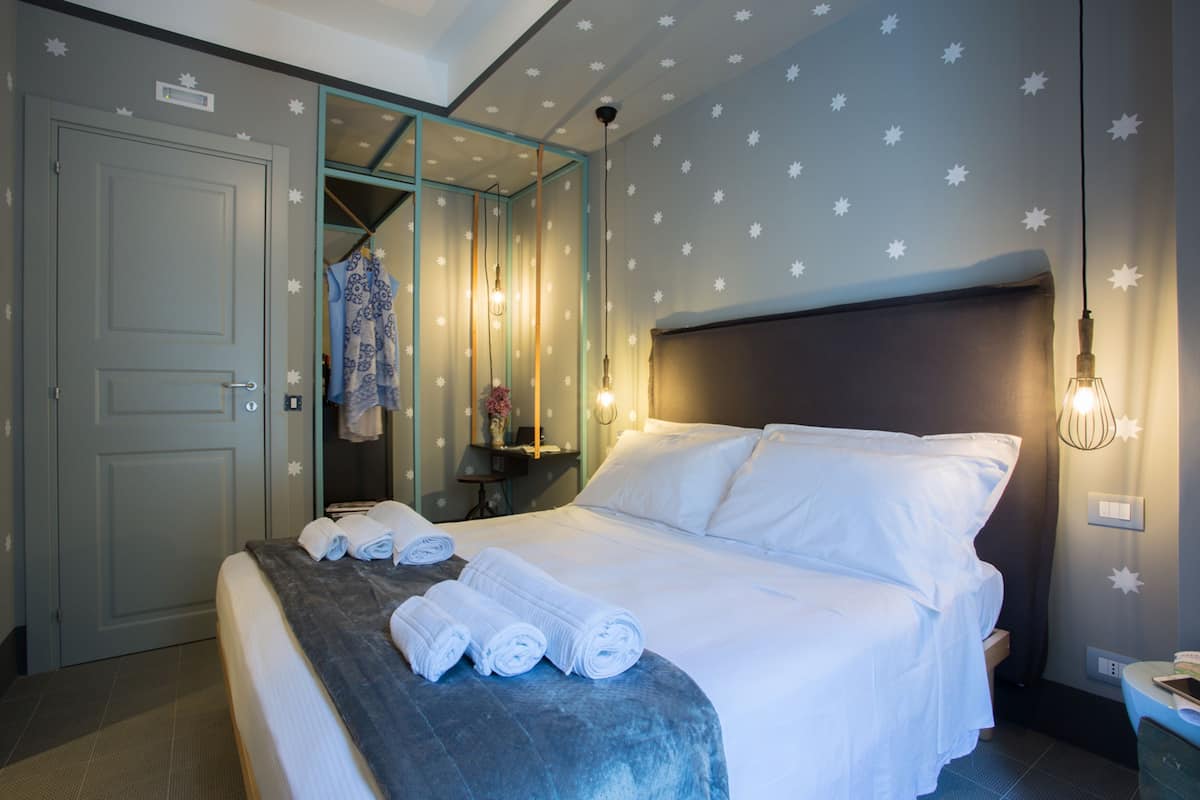 A hotel room with a bed, a bathroom with a shower curtain, and a nightstand with a lamp.