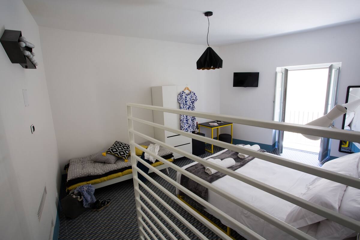 A compact hotel room with a single bed, a small desk, a chair, a hanging light fixture, and a window providing natural light.