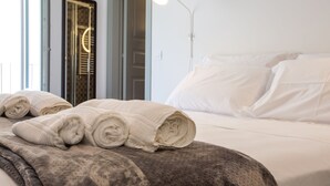 Una cama impecable con toallas enrolladas, una lámpara de noche y vista a una puerta con un espejo circular.